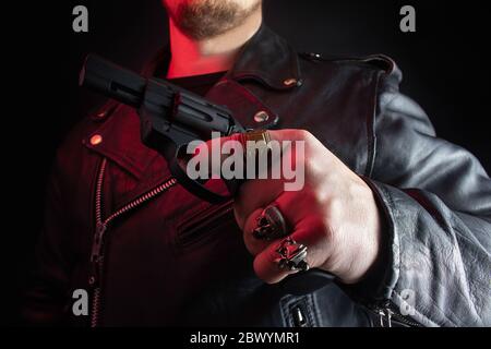 Foto eines Biker in schwarzer Lederjacke mit Totenkopf Ringe halten Revolver auf schwarzem Hintergrund. Stockfoto