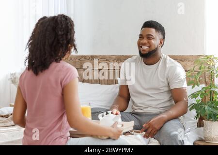 Fröhlicher schwarzer Mann, der seine Freundin anschaut, Frühstück im Bett Stockfoto