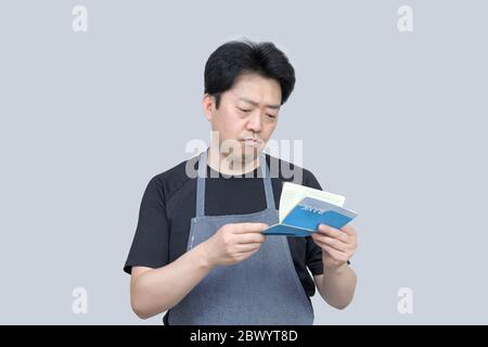Ein asiatischer Mann mittleren Alters, der ein Bankpassbuch in der Hand auf grauem Hintergrund hält. Stockfoto