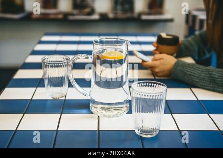 Ein Dekanter und ein Glas Wasser mit Zitrone im Vordergrund. Im Hintergrund benutzt das Mädchen ein Mobiltelefon. Sie hat ein Treffen oder sie ruht sich aus und trinkt Kaffee. Stockfoto