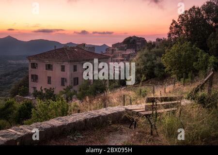 Eine alte Bank mit Blick auf den Sonnenuntergang über dem alten Bergdorf Belgodere in der Balagne von Korsika mit dem Mittelmeer in der Stockfoto