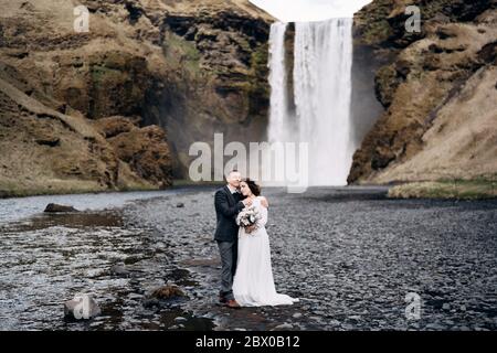 Hochzeit in Island. Hochzeitspaar in der Nähe des Skogafoss Wasserfalls. Die Braut und der Bräutigam umarmen sich am Fluss. Stockfoto