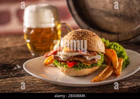 Hühnerburger mit Spiegelei und Speck, Salat, Paprika und Kartoffelkeile auf einem Teller neben einem Bier vom Fass serviert Stockfoto