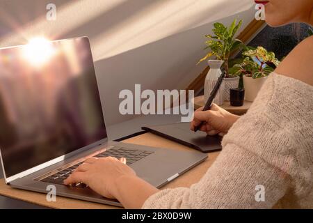 Eine kaukasische Frau mit rotem Lippenstift arbeitet an ihrem Laptop, während sie während eines goldenen Sonnenuntergangs ein Grafiktablett benutzt Stockfoto