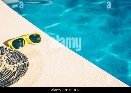 Gelbe Plastiksonnenbrille und ein Strohhut auf einem Stein am Wasser sind hellblau - ein heißer Sommertag Stockfoto