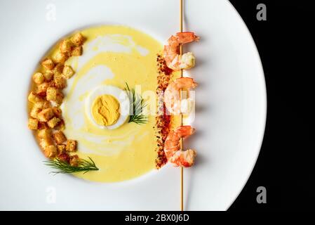 Warme Sahnesuppe mit Käse, Ei, frischem grünem Dill, Garnelen und Gewürzen, gebratenen Croutons, auf einem weißen runden Teller auf schwarzem Hintergrund Stockfoto