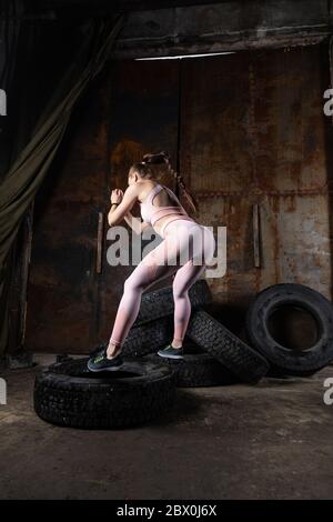 Sportliche Frau Trainer springt auf einen Reifen, macht Fitness in einer alten Garage. Das Konzept des Sports unter schwierigen unangemessenen Bedingungen, ersetzt Spec Stockfoto