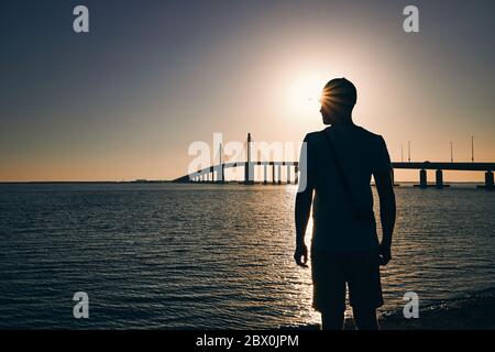 Silhouette des jungen Mannes am Strand gegen lange Brücke über das Meer bei Sonnenuntergang. Abu Dhabi, Vereinigte Arabische Emirate Stockfoto