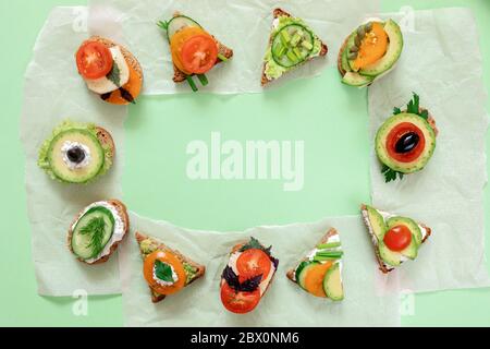 Rahmen aus vegetarischen Sandwiches mit Tomaten, Gurken, Avocado, sonnengetrockneten Oliven, Grünes und Käse im Kreis auf Pergamentpapierstücken gelegt Stockfoto