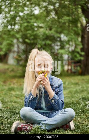 Schönes Mädchen mit blonden Haaren zog ihre Maske aus und schnüffelt Löwenzahn, der Frühling kommt Stockfoto