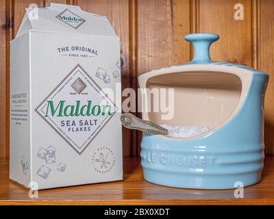 Packung Maldoner Salzflocken neben einem hellblauen Keramiksteinzeug, Le Creuset Salzschwein, hält einen kleinen Löffel in Salzflocken, in einem Pinienschrank. Stockfoto