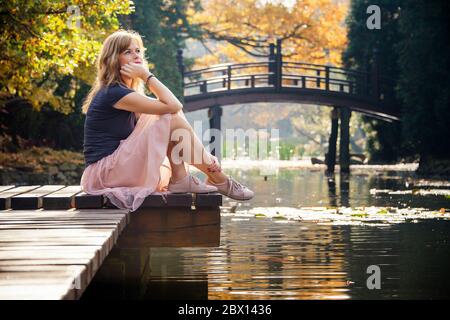 Träumende junge Frau sitzt auf einem hölzernen Pier in der Nähe von See im Herbst Stadtpark Stockfoto