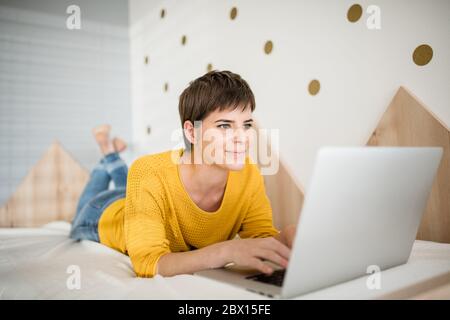 Junge Frau mit Laptop liegt auf dem Bett im Schlafzimmer drinnen zu Hause. Stockfoto