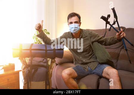 Maskierter Mann bereit für Abenteuer mit Rucksack und Accessoires in seinem Wohnzimmer während der Coronavirus-Saison vorbereitet Stockfoto