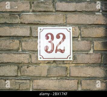 Hausnummer 32 zweiunddreißig. Braune Zahlen auf einer weißen Platte, die zu einer alten Ziegelmauer gebaut wurde Stockfoto