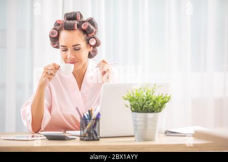 Frau mit Haarwalzen und Nachthemd riecht Kaffee, während sie zu Hause vor dem Computer sitzt. Stockfoto