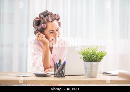 Weibchen in Haarwalzen und Nachthemd lacht über Notebook-Scrren, während sittinf am Tisch zu Hause. Stockfoto