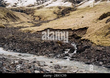 Gebirgsfluss am Fuße des Berges mit einem schneebedeckten Gipfel. Gelbes trockenes Gras auf den Bergen im Mai in Island. Stockfoto