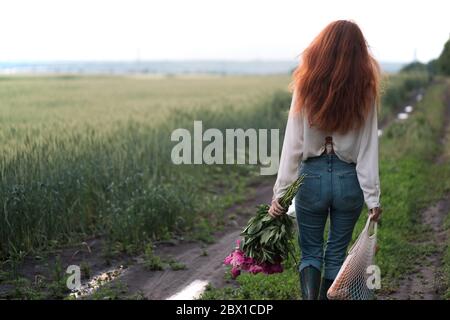Ein Mädchen mit einem Blumenstrauß und einem Saitenbeutel mit Baguettes geht auf einer Landstraße. Frühlingsstimmung im Dorf. Stockfoto