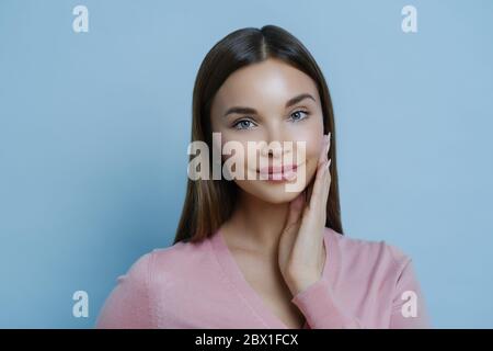 Foto von entzückenden jungen Millennial weiblich berührt Wange sanft, genießt Weichheit der Haut nach der Anwendung Schönheitsmaske oder Gesichtscreme, blickt auf Kamera, iso Stockfoto