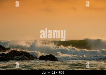 Wellen brechen an einer felsigen Küste bei Sonnenaufgang. Goldenes Leuchten spiegelt sich über dem glitzernden Wasser. Stockfoto