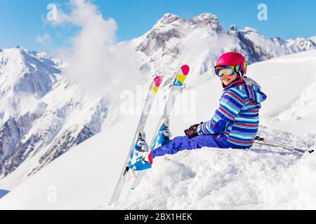 Niedliches Kind im hellen Sport-Outfit sitzen auf dem Schneehaufen im Berg über hohe Gipfel Blick zurück zur Kamera Stockfoto