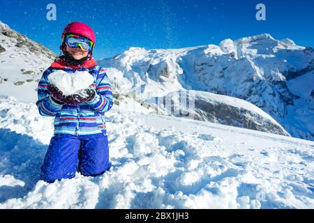 Lächelnd glücklich niedliches Mädchen in Ski-Outfit Maske und Helm zeigen Liebe Konzept hält Schnee Form im Herzen auf den Knien mit Bergen auf dem Rücken stehen Stockfoto