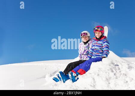 Zwei schöne Mädchen sitzen auf Haufen Schnee über blauem Hintergrund in Himmel Outfit Farbe Maske und Brille Blick auf Kamera lächelnd Stockfoto