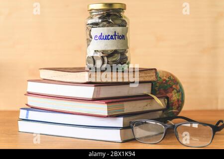 Münzen im Glas mit Bildungsetikett, Bücher, Gläser und Globus auf Holztisch. Finanzkonzept. Stockfoto