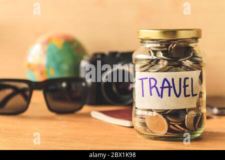 Münzen in Glas mit Accessoires des Reisenden auf Holztisch. Geld sparen für Reisekonzept Stockfoto