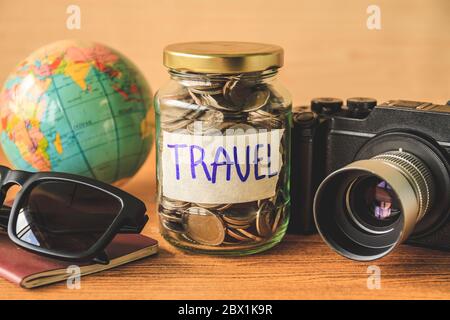 Münzen in Glas mit Accessoires des Reisenden auf Holztisch. Geld sparen für Reisekonzept Stockfoto