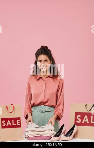 Porträt einer jungen hübschen Frau mit lockigen Haaren lächelnd vor der Kamera, während sie in der Nähe der Einkaufstaschen mit neuen Kleidern steht Stockfoto