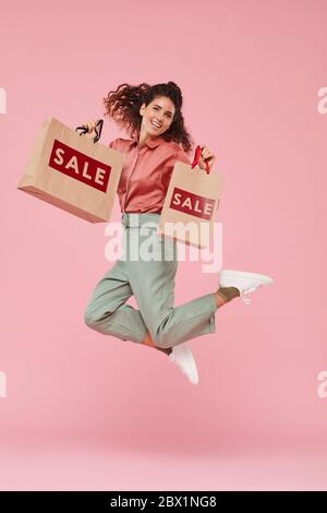 Porträt der jungen Frau glücklich mit ihrem erfolgreichen Einkauf sie springt vor dem rosa Hintergrund Stockfoto