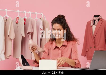 Junge lächelnde Frau sitzt am Tisch und untersucht ihre neuen Schuhe, die sie online bestellt Stockfoto