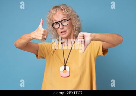 Ältere Frau mit Brille zeigt Daumen nach oben und Daumen nach unten, schwierig zu wählen Stockfoto