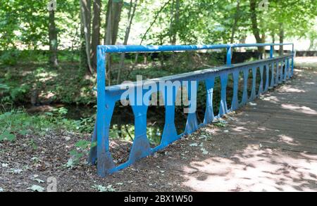 Alte blaue Metallbrücke mit Holzplanken über einem kleinen Fluss in einem Park mit viel Grün, Bäumen und Wanderweg Stockfoto