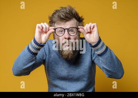 Seltsamer, zotteliger, bärtiger Mann in einem alten Pullover hält Gläser in den Händen und blinkend, er sah etwas Interessantes und war überrascht. Stockfoto