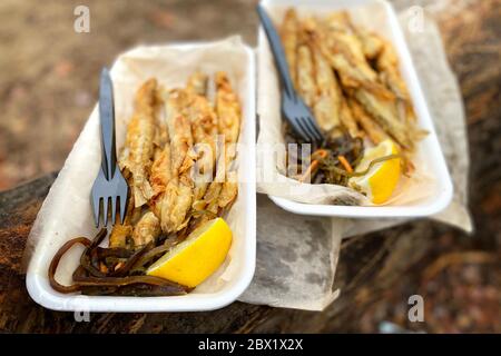 Gebratene roch auf Stück Backpapier auf braunem Holz in auf einem Picknick im Wald, Nahaufnahme. Kleiner Fisch neben einer Zitrone und Seekohl. Blick von oben. Stockfoto