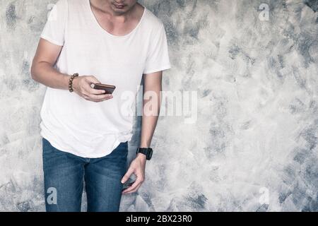 Junger Mann, der weißes T-Shirt und blaue Jeans trägt, Smartphone hält und auf grauem Grunge-Hintergrund steht. Freier Platz für Text Stockfoto