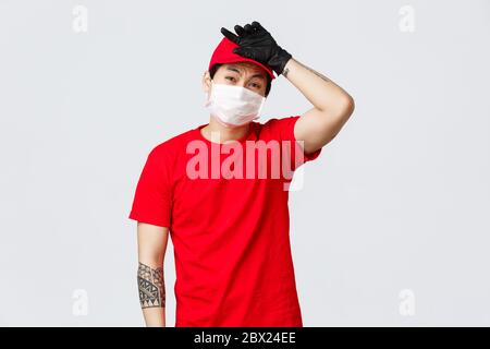 Puh harte Arbeit der Kuriere. Müde asiatische Lieferung Kerl in medizinischen Maske und Handschuhe, tragen rote Kappe und T-Shirt Uniform, seufzen, wischen Schweiß von der Stirn Stockfoto