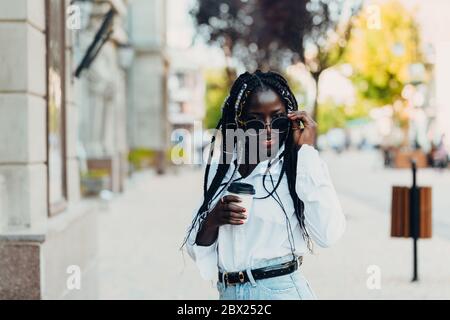 Porträt eines lächelnden jungen dunkelhäutigen Mädchens mit Zöpfen in Gläsern mit Kaffee in der Hand im Freien an einem sonnigen Tag. Außenfoto. Stockfoto