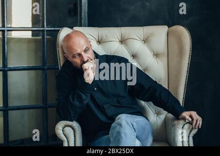 Trauriger, ernster Mann mittleren Alters in einem schwarzen Hemd sitzt und denkt über seine Probleme in einem Raum mit dunklen Wänden nach Stockfoto