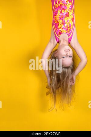 Aufgeregt verrückte kleine blonde Mädchen in einem Badeanzug, hängend kopfüber auf einem gelben Studio Hintergrund. Emotion, Ausdruck. Platz für Text kopieren Stockfoto