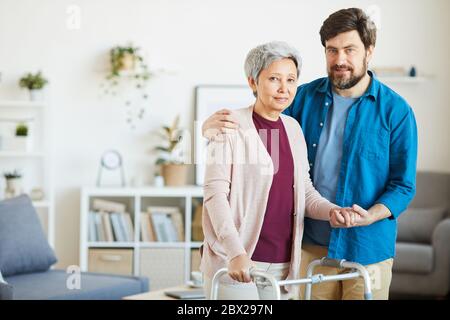 Porträt des Sohnes umarmt seine ältere Mutter mit Wanderer, während sie im häuslichen Raum stehen und Blick auf die Kamera Stockfoto