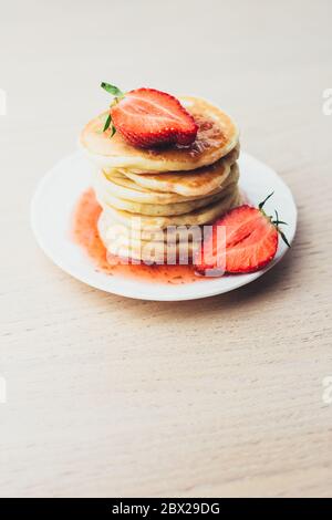 Vertikales Bildformat - ein Stapel von frisch duftenden heißen Pfannkuchen auf einem weißen Teller mit Erdbeermarmelade gegossen - frische Erdbeere Stockfoto