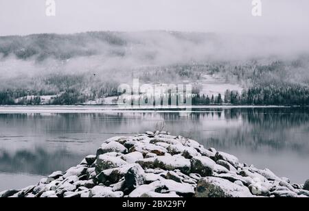 Schneebedeckte Felsen auf einem See mit Hügeln und Wald in Nordschweden. Stockfoto