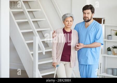 Porträt der männlichen Krankenschwester in Uniform hält für die Hand der älteren Frau und sie schauen auf Kamera, während zu Hause stehen Stockfoto