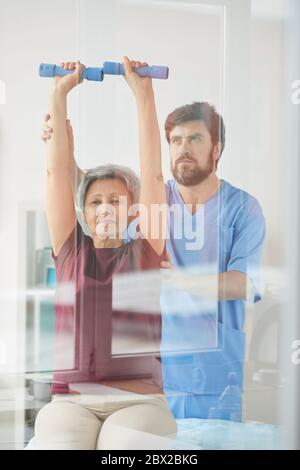 Ältere Frau, die ihre Hände hochhebt und mit Hanteln trainiert, mit einem Arzt, der ihr im Krankenhaus hilft Stockfoto