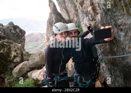 Konzept: Abenteuer. Paar Kletterer mit Helm und Geschirr. Aufnehmen eines Selfies mit dem Smartphone. Auf dem Gipfel des Berges. Klettersteig in r Stockfoto