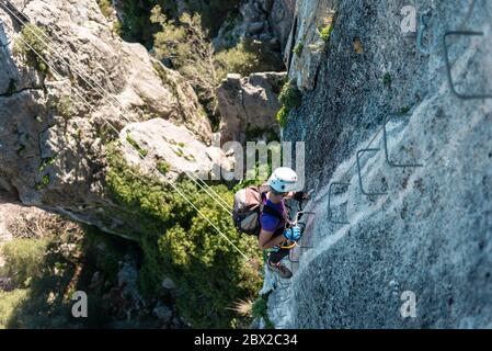 Konzept: Abenteuer. Frau mit Helm, Geschirr und Rucksack. Gehen Sie die eiserne Treppe hinunter, die an die Wand geheftet ist. Klettersteig in den Bergen. Stockfoto